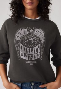 Sötétszürke pulóver, amelyen nagyméretű grafikai dizájn található fehér szöveggel, amely szárnyakat és a "QUALITY CLOTHING SAN FRANCISCO CAL" kifejezést ábrázolja.