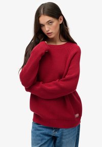 Maglione rosso lavorato a maglia con maniche lunghe, scollo rotondo e polsini e orlo a costine. Presenta una piccola patch con logo sul lato inferiore.