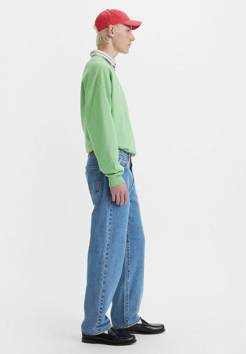 Felpa verde oversize, colletto a righe, jeans baggy azzurri, scarpe nere e cappellino rosso; texture liscia, design casual.