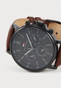 Reloj cronógrafo negro con correa de cuero marrón. Presenta un dial oscuro, marcadores de hora plateados, displays de fecha y día, y subdiales.