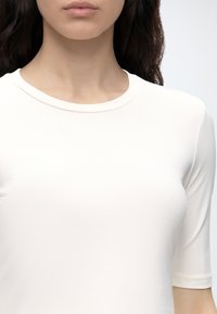 Femme portant un t-shirt blanc uni à manches courtes et col rond, montrant le cou et le haut de la poitrine, avec les cheveux foncés partiellement visibles.