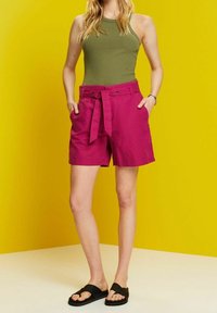 Shorts fucsias con un lazo en la cintura, bolsillos laterales y un corte relajado, combinados con una camiseta sin mangas verde y sandalias negras, contra un fondo amarillo.