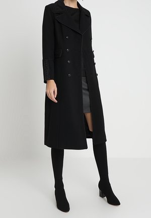 Osoba ubrana w długi czarny dwurzędowy płaszcz, czarną spódnicę i czarne botki na obcasie, stojąca na jednolitym tle.