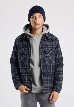 Laivastonsininen ruutukuosinen takki, jossa kaksi rintataskua, valmistettu teksturoidusta kankaasta, kerroksittain vaaleanharmaan hupullisen paidan päällä ja yhdistetty harmaisiin farkkuihin.