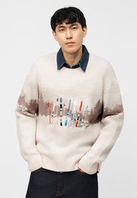 Νέος άνδρας που φοράει μπεζ πουλόβερ με πολύχρωμο σχέδιο από μπαστούνι σκι και καφέ σχέδιο δάσους, σκούρο τζιν και μπλε πουκάμισο από denim από κάτω.