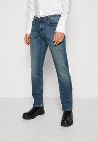 Blue denim jeans with a straight cut, featuring subtle fading, classic five-pocket design, and a button closure. Paired with black ankle boots.