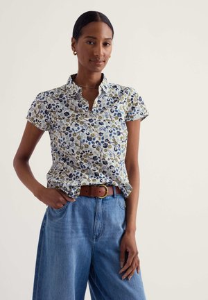 Tumejuustega naine, kes kannab kuldseid rõngaskõrvarõngaid, lühikese varrukaga lillelist nööbitavat särk, pruun vöö ja sinised kõrge vöökohaga teksad.