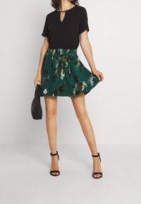 Schwarzes kurzärmliges Oberteil mit einem Schlüsselloch-Ausschnitt, kombiniert mit einem grünen Rock im Blumenmuster. Das Model hält eine schwarze geflochtene Tasche und trägt schwarze Absatzsandalen.