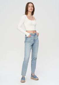 Biały prążkowany krótki top z długim rękawem i detalem guzika, zestawiony z jasnoniebieskimi dżinsami oraz masywnymi niebieskimi butami wsuwanymi.