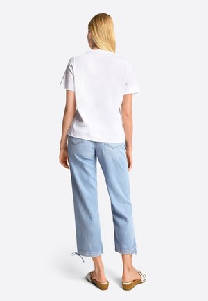 Kvinde med langt blondt hår iført en simpel hvid t-shirt, lyseblå cropped jeans med påtie ved kanten og hvide hæle sandaler, stående med ryggen til.