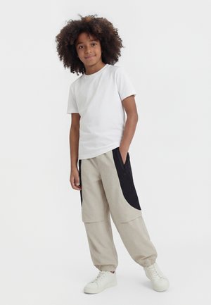 Παιδί με κατσαρά μαλλιά που φοράει λευκό μπλουζάκι, μπεζ και μαύρο παντελόνι cargo και άσπρα αθλητικά παπούτσια, στέκεται με το ένα χέρι στην τσέπη.