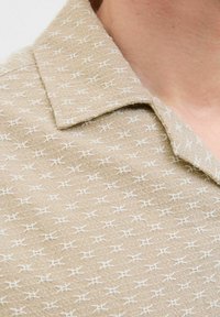Camicia beige con colletto realizzata in tessuto texturizzato, caratterizzata da un motivo geometrico bianco ripetitivo, che esalta l'aspetto intrecciato.