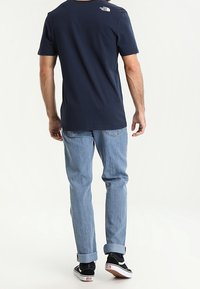 Námořnická modrá bavlněná tričko s kulatým výstřihem, světle modré džíny a černé tenisky s bílou podrážkou, pohled zezadu.