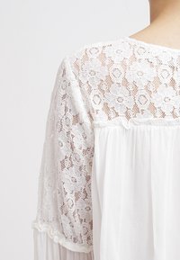 Blouse blanche avec empiècement en dentelle florale et tissu translucide fluide. Présente un léger froncé à la taille et une délicate bordure en dentelle aux manches.