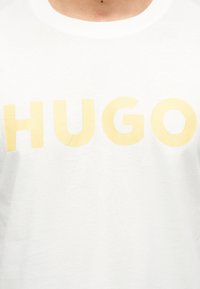 Λευκό Τ-shirt με μεγάλα, ανοιχτόκκινα κεφαλαία γράμματα που σχηματίζουν τη λέξη "HUGO" στο στήθος ενός ατόμου που το φοράει.