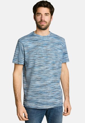 Homme portant un t-shirt à manches courtes bleu et gris à motifs et un jean bleu, debout devant un fond clair uni.