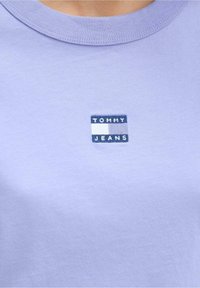 Světle fialové bavlněné tričko s obdélníkovým vyšívaným logem v námořnické modři a bílé barvě, s nápisem "TOMMY JEANS" uprostřed na hrudi.