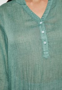 Blusa di lino teal con scollatura a V, tre bottoni e una finitura testurizzata. Presenta un leggero luccichio e un tessuto leggero.