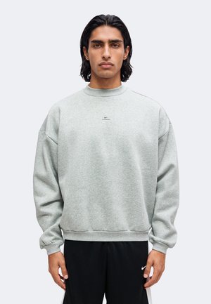 Homme aux cheveux bruns aux épaules, portant un sweat-shirt gris clair et un pantalon noir, debout devant un fond uni.