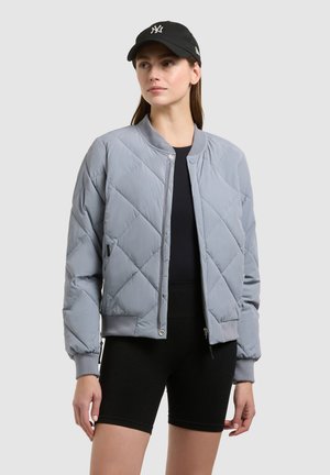 Ženska, oblečena v svetlo sivo prešito bomber jakno, črne kratke hlače, črno majico in črno kapo z belim logotipom, stoji pred enobarvnim ozadjem.