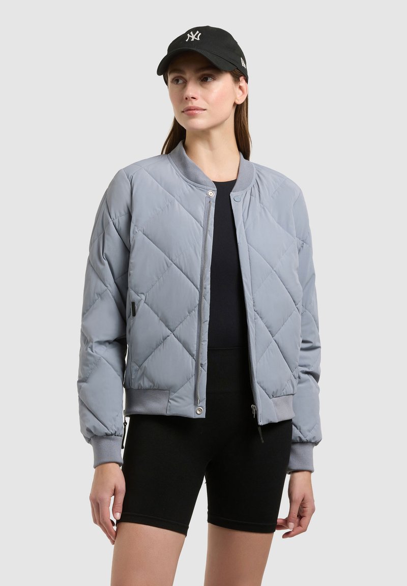 Femme portant une veste bomber matelassée gris clair, un short noir, un haut noir et une casquette noire avec un logo blanc, se tenant devant un fond uni.