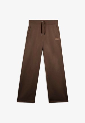 Pantalons de survêtement marron à jambes larges en tissu doux. Dotés d'une taille élastique avec cordon de serrage et de poches latérales. Marquage subtil sur la hanche.