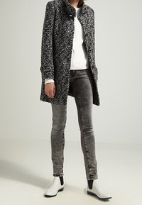 Uldblandet frakke i sort og hvid houndstooth-mønster med høj krave, to knapper, parret med grå jeans og hvide ankelstøvler.