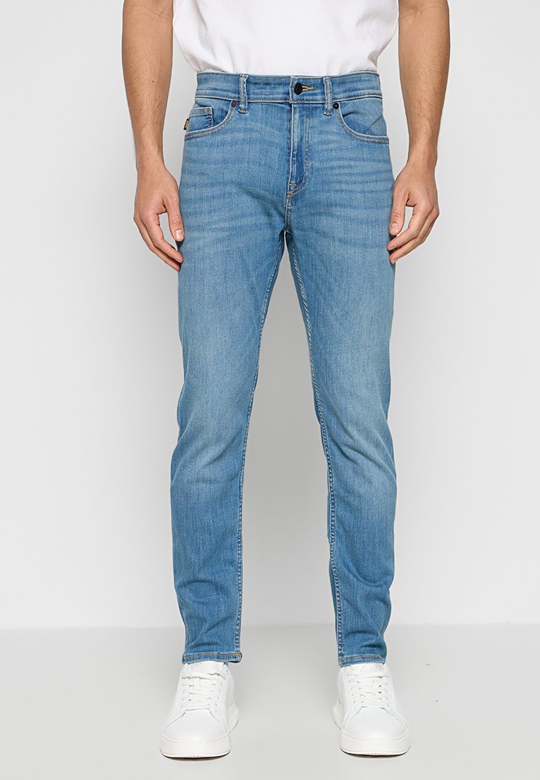 Jean en denim bleu clair avec une coupe droite, design à cinq poches, décoloration subtile et coutures contrastées. Porté avec une chemise blanche et des baskets.