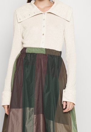 Kobieta ubrana w jasnobeżową bluzkę z guzikami, szerokim kołnierzem i długimi mankietami, w połączeniu z wysokim stanem i patchworkową spódnicą w odcieniach zieleni i brązu.
