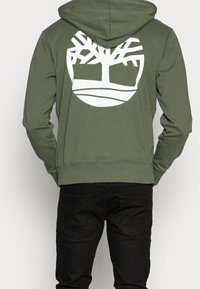 Sudadera verde con un logo de árbol blanco en la parte trasera, hecha de una mezcla de algodón, con capucha ajustable y dobladillo acanalado. Combinada con jeans negros.
