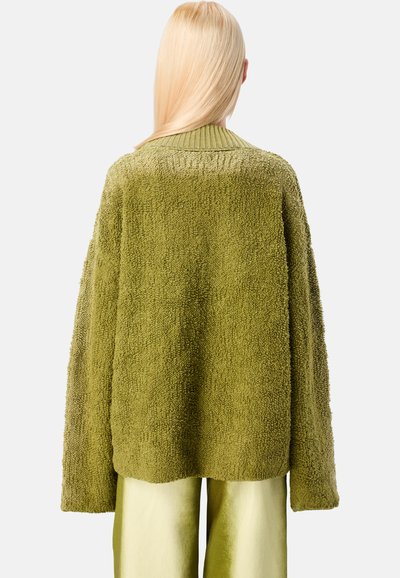 Henkilö, jolla on pitkät vaaleat hiukset, pukeutuu löyhään, teksturoituun oliivinvihreään neuleeseen ja kiiltäviin vaaleanvihreisiin housuihin, nähtynä selkäpuolelta.