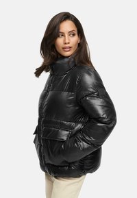 Black puffer jacket with a shiny, quilted exterior, high collar, and two front pockets. Features a small brand patch on the sleeve.