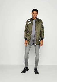 Joven de pie, vistiendo una chaqueta bomber verde oliva con parches, camiseta de rayas, jeans ajustados grises y botines negros.