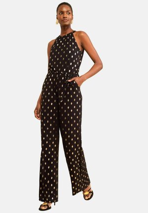 Frau trägt einen ärmellosen schwarzen Jumpsuit mit metallischem goldfarbenem Punktmuster, Seitentaschen, gebundener Taille und schwarzen Sandalen mit goldenen Akzenten.