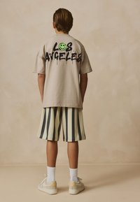 Beige übergroßes T-Shirt mit schwarzem Graffiti-Schriftzug "LOS ANGELES" und einem grünen Smiley. Kombiniert mit gestreiften Shorts in Hellgrün, Schwarz und Grau.