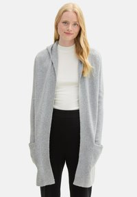 Ljusgrå cardigan med huva, ribbad textur och öppen fram. Har två framfickor och en avslappnad passform, stiliserad över en vit skjorta.