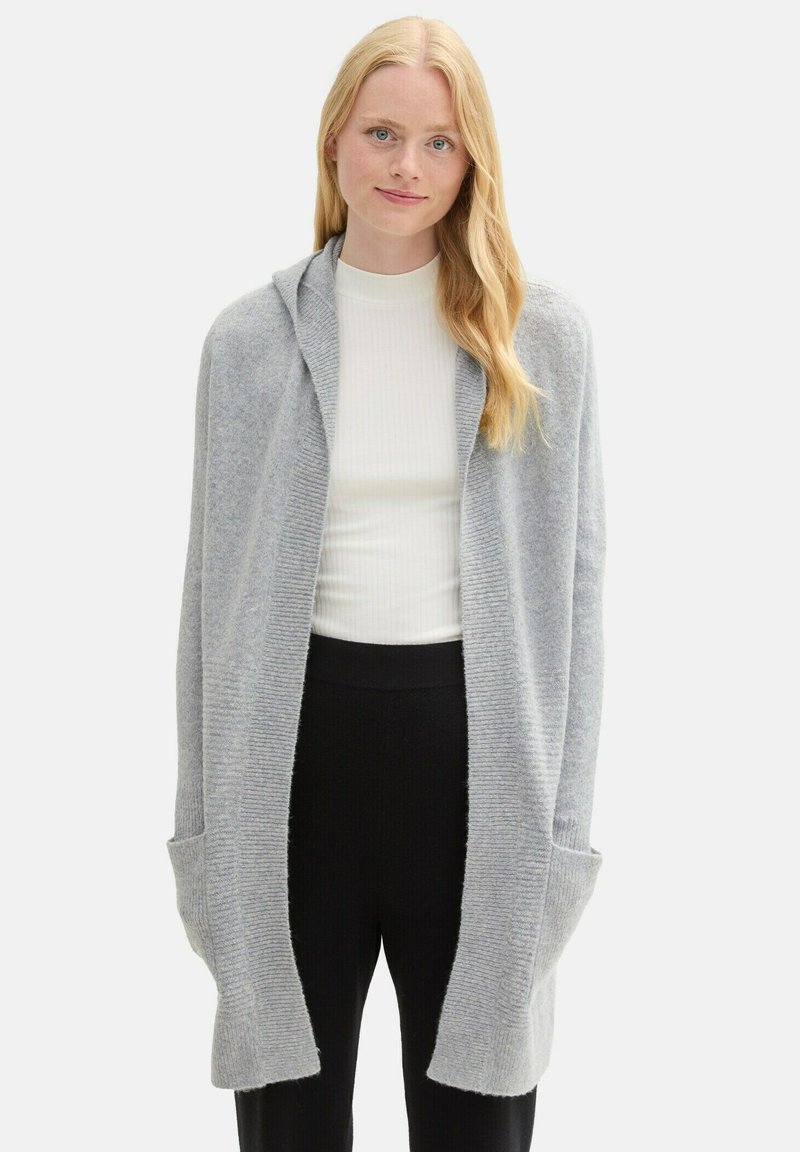 Ljusgrå cardigan med huva, ribbad textur och öppen fram. Har två framfickor och en avslappnad passform, stiliserad över en vit skjorta.