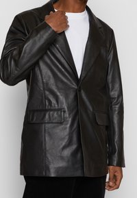 Svart läderblazer med sned krage, enkel knappstängning och två framfickor. Slät textur med synliga sömdetaljer.