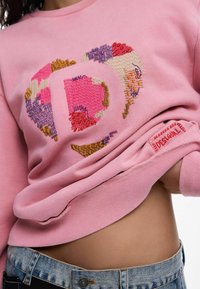 Rožinė džemperis su spalvingu siuvinėtu širdies motyvu. Turi ribotus rankogalius, logotipo žymę apačioje ir laisvą siluetą.