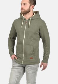 Olivgrüner Zip-Hoodie aus Baumwollmischung, mit Kapuze inklusive Kordelzug, Fronttaschen und einem Logo-Patch aus Kunstleder.