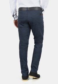 Man draagt marineblauwe slim-fit broek, zwarte riem, wit overhemd en zwarte schoenen, staat met de rug naar voren met handen ontspannen langs zijden.
