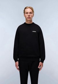 Giovane con capelli biondi corti, che indossa una felpa nera e pantaloni neri, in piedi davanti a uno sfondo grigio chiaro uniforme.