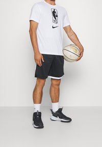 T-shirt blanc avec logo NBA noir, short noir avec des accents blancs et baskets noires. Tenant un ballon de basket avec des détails dorés.