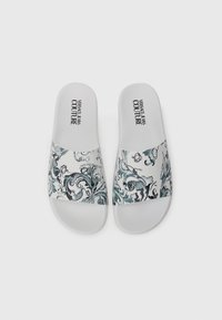 White slide sandals featuring a swirling green and black pattern on the strap. The footbed is textured, and “Versace Jeans Couture” is printed on top.