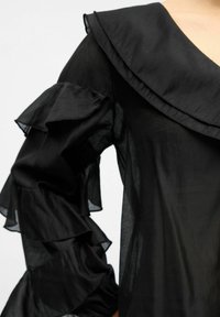 Blouse noire transparente avec des manches à volants superposées et un large col pointu, portée sur le haut du torse devant un fond uni.