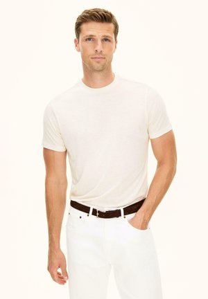 Junger Mann mit kurzen blonden Haaren, trägt ein schlichtes weißes T-Shirt und weiße Jeans, eine Hand in der Tasche, blickt nach vorne vor hellem Hintergrund.