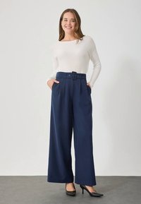Μπλε σκούρο παντελόνι με φαρδιά γραμμή, ψηλή μέση με ζώνη, πλαϊνές τσέπες και λείο φινίρισμα υφάσματος. Συνδυασμένο με ανοιχτόχρωμο μακρυμάνικο τοπ και τακούνια.