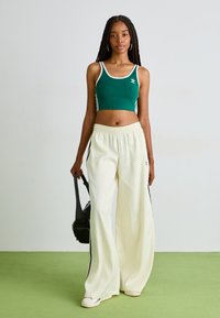 Top cropped verde com acabamento branco, combinado com calças brancas largas com riscas laterais pretas, modelo a segurar uma bolsa preta.