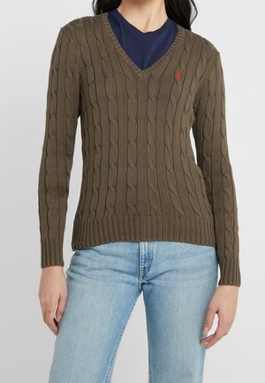 Pull en maille torsadée vert olive avec col en V et poignets côtelés. Présente un petit logo sur la poitrine. Associé à un jean en denim bleu clair.