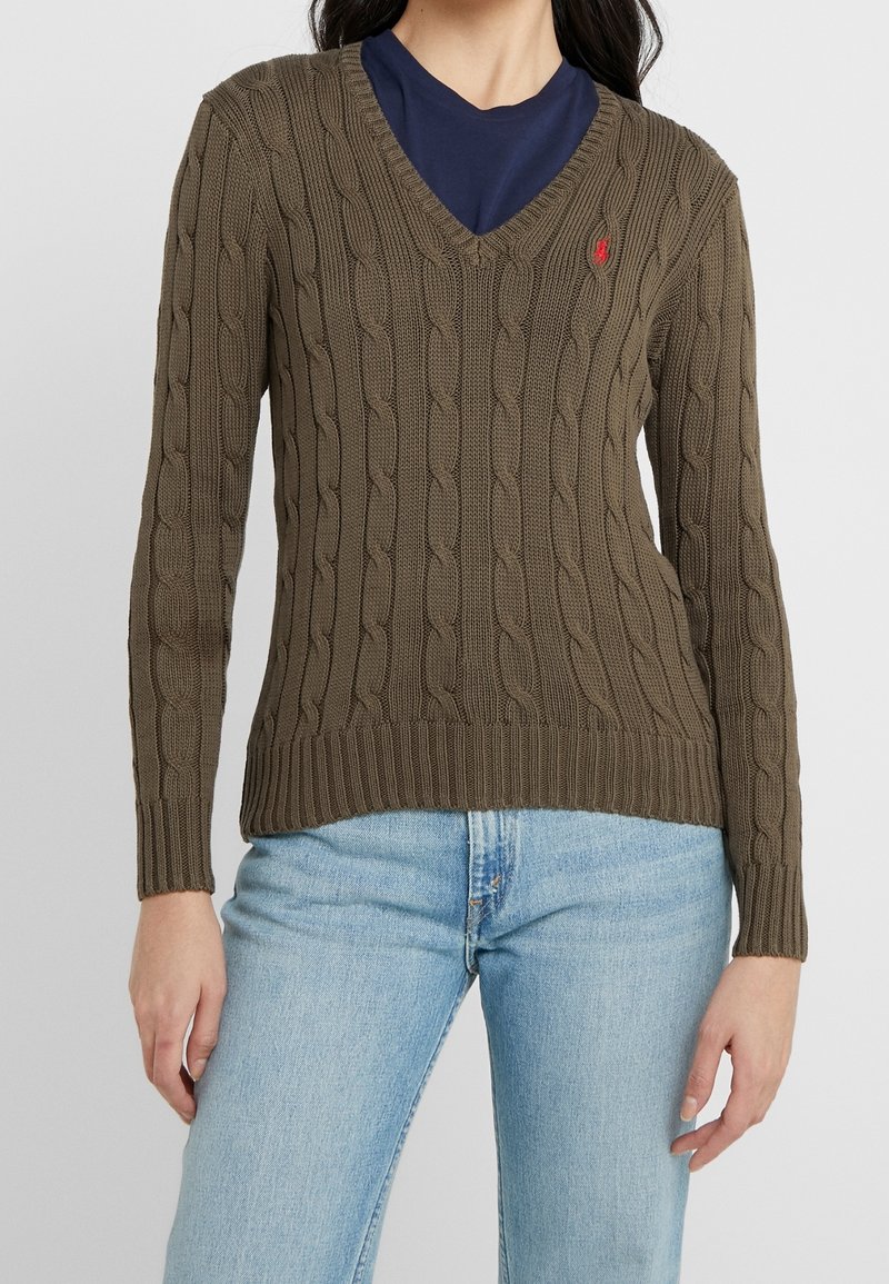 Pull en maille torsadée vert olive avec col en V et poignets côtelés. Présente un petit logo sur la poitrine. Associé à un jean en denim bleu clair.